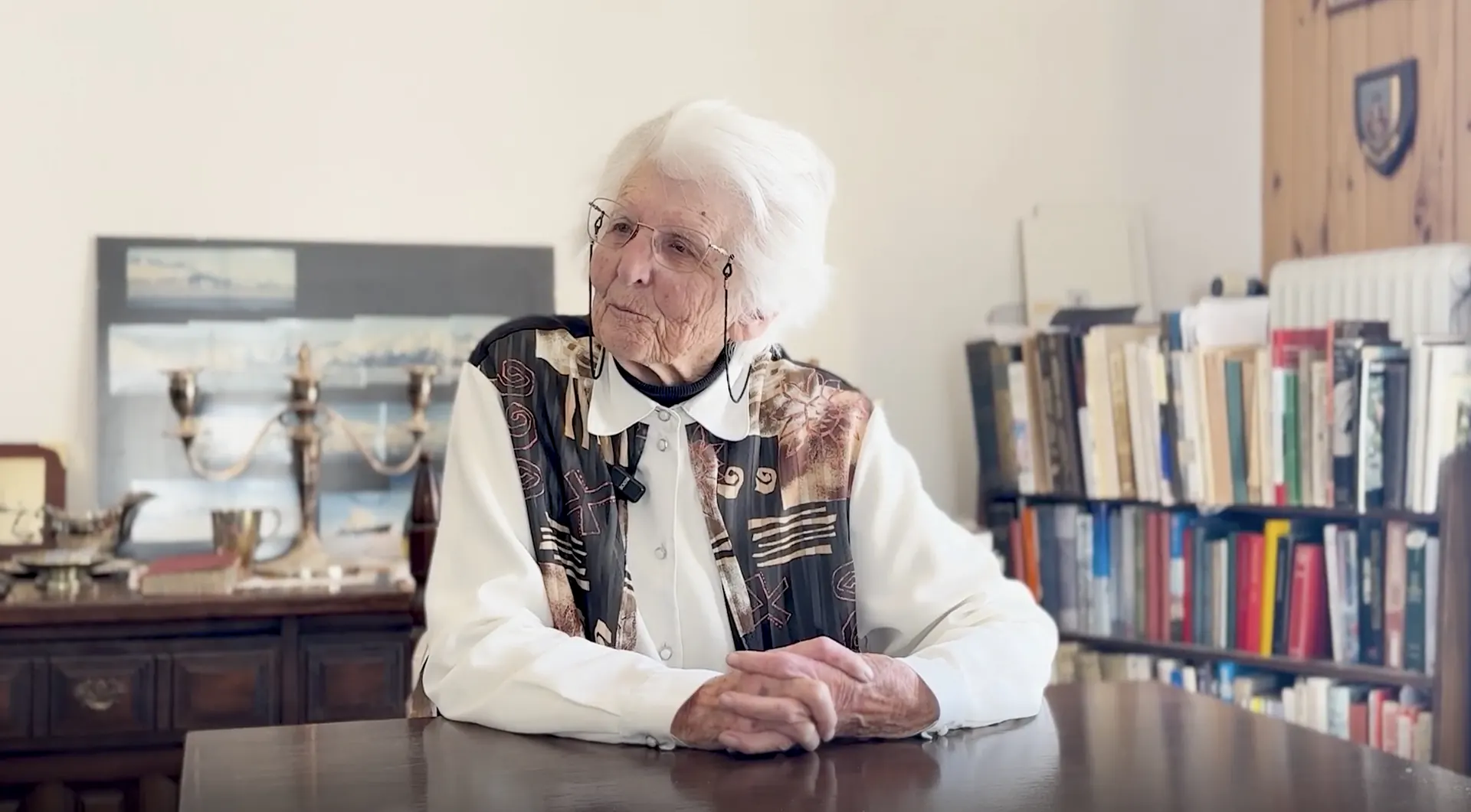 Madame Joanne seated at a wooden desk in a book-lined room, speaking to camera with a microphone clip