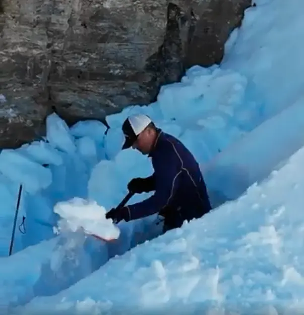 A person in a helmet and dark jacket chipping blue glacial ice with a tool on a steep glacier face