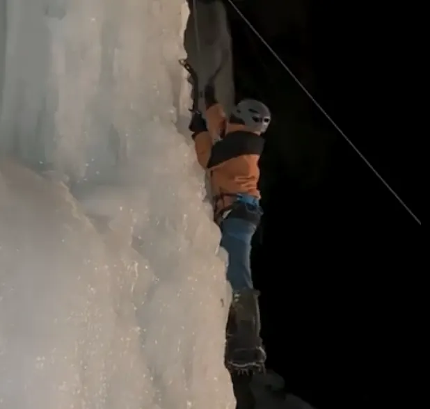 Climber in orange jacket ascending a frozen waterfall with ice axes and crampons