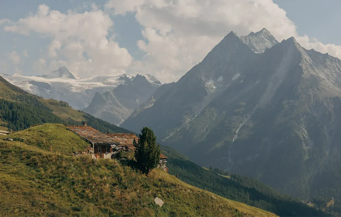 Stone alpine chalet on a grassy ridge facing snow-capped mountain peaks