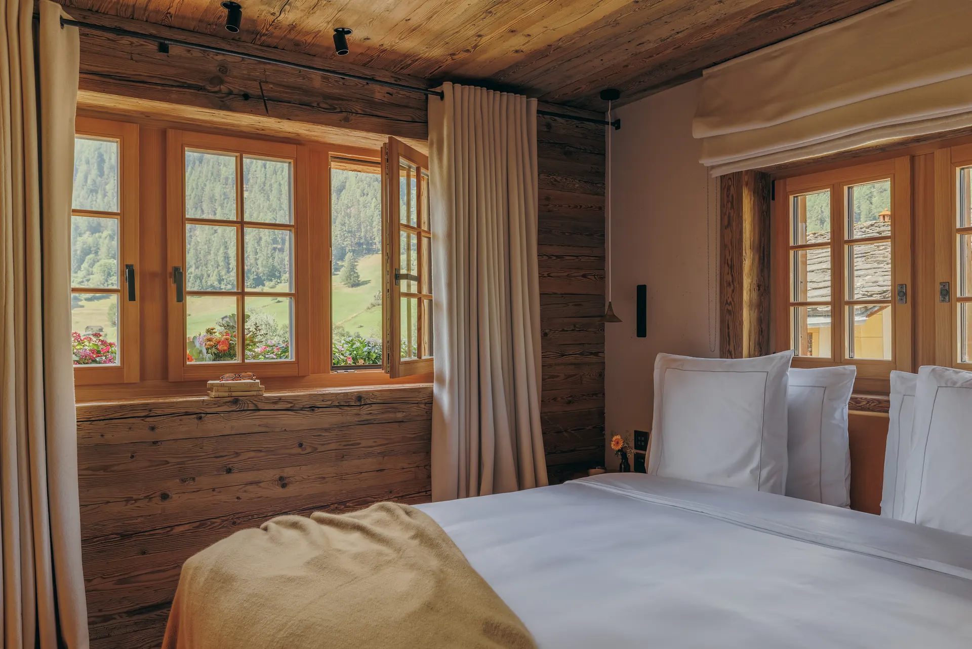 White bed in a wood-paneled corner between two windows, one open onto a green mountain valley