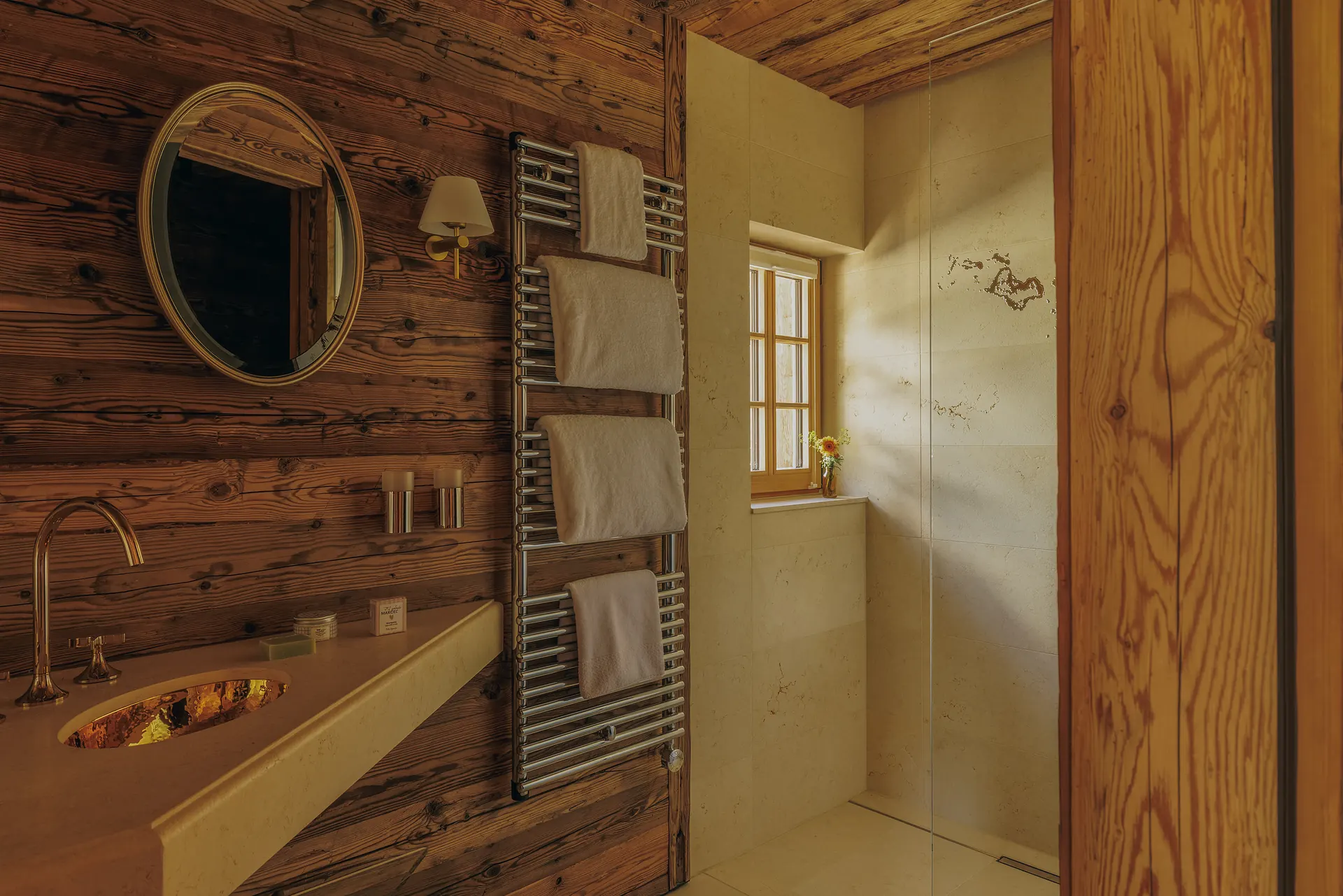 Bathroom with a round gold mirror, heated towel rail and glass-screened shower lit by a window