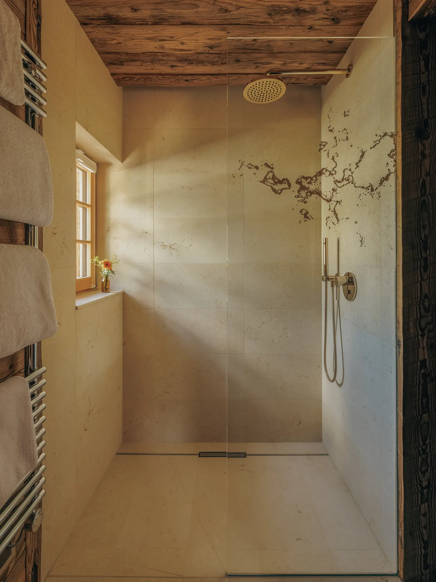 Walk-in shower with pale stone walls, brass rain shower head and sunlight from a small window