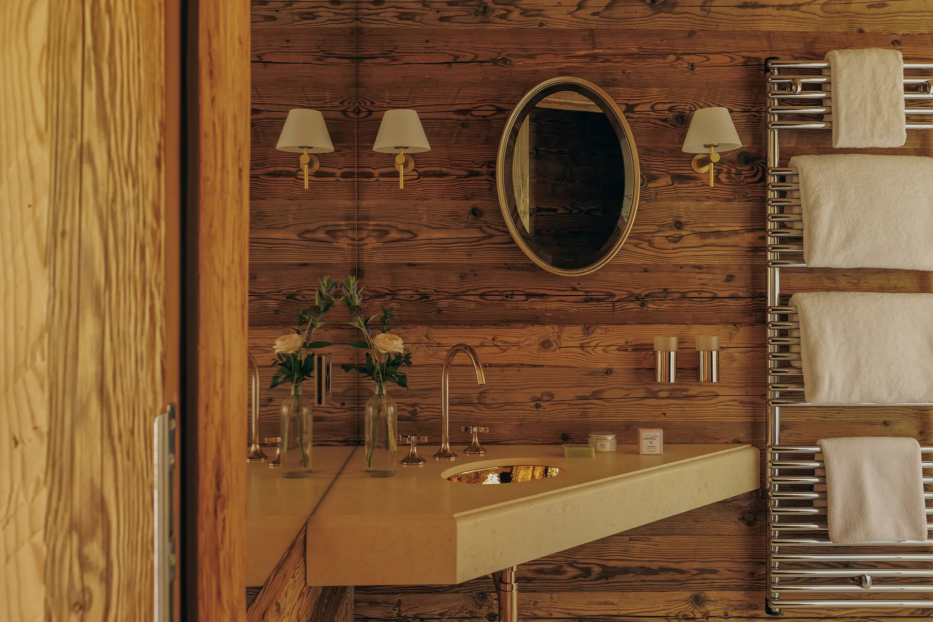 Stone vanity with a hammered brass basin, gold oval mirror and wall sconces on weathered wood