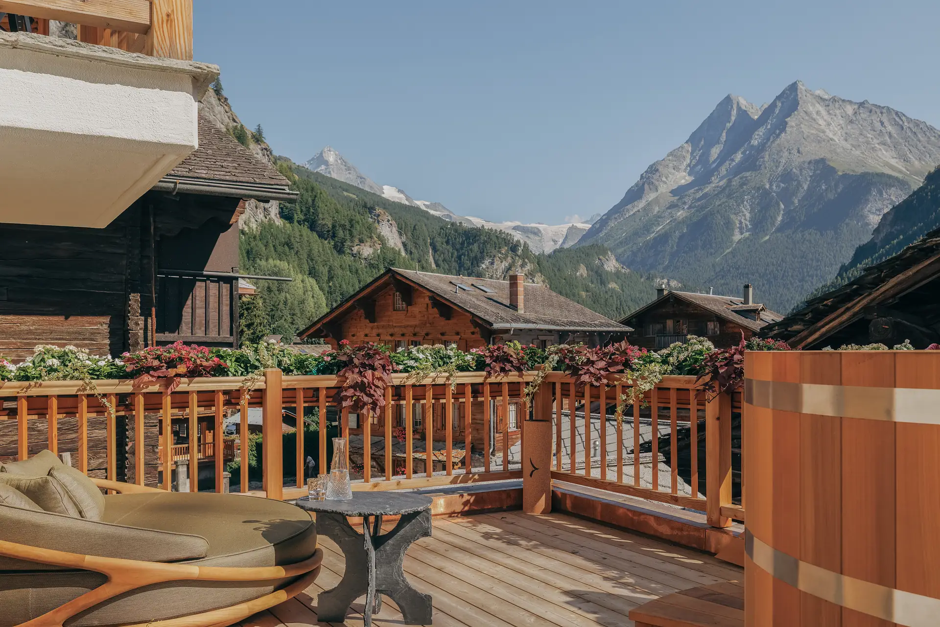 Wooden terrace with a lounge daybed, slate side table and wooden hot tub facing snowy alpine peaks