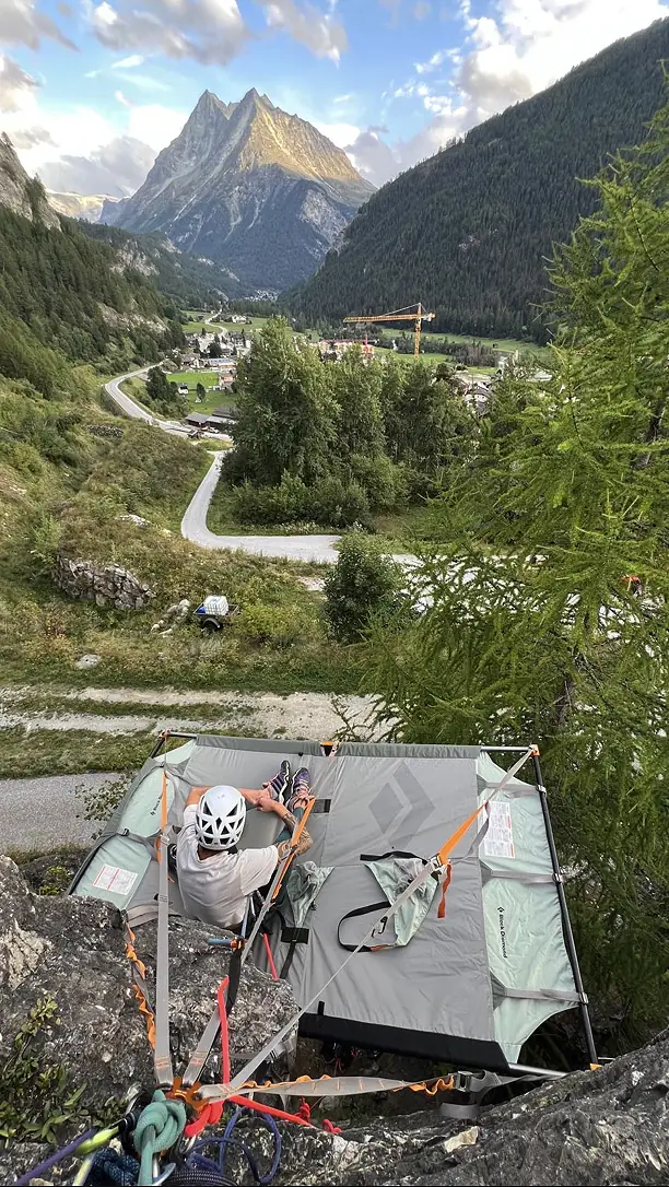 Climber in a white helmet resting on a portaledge hung from a cliff above a mountain village and pointed peak