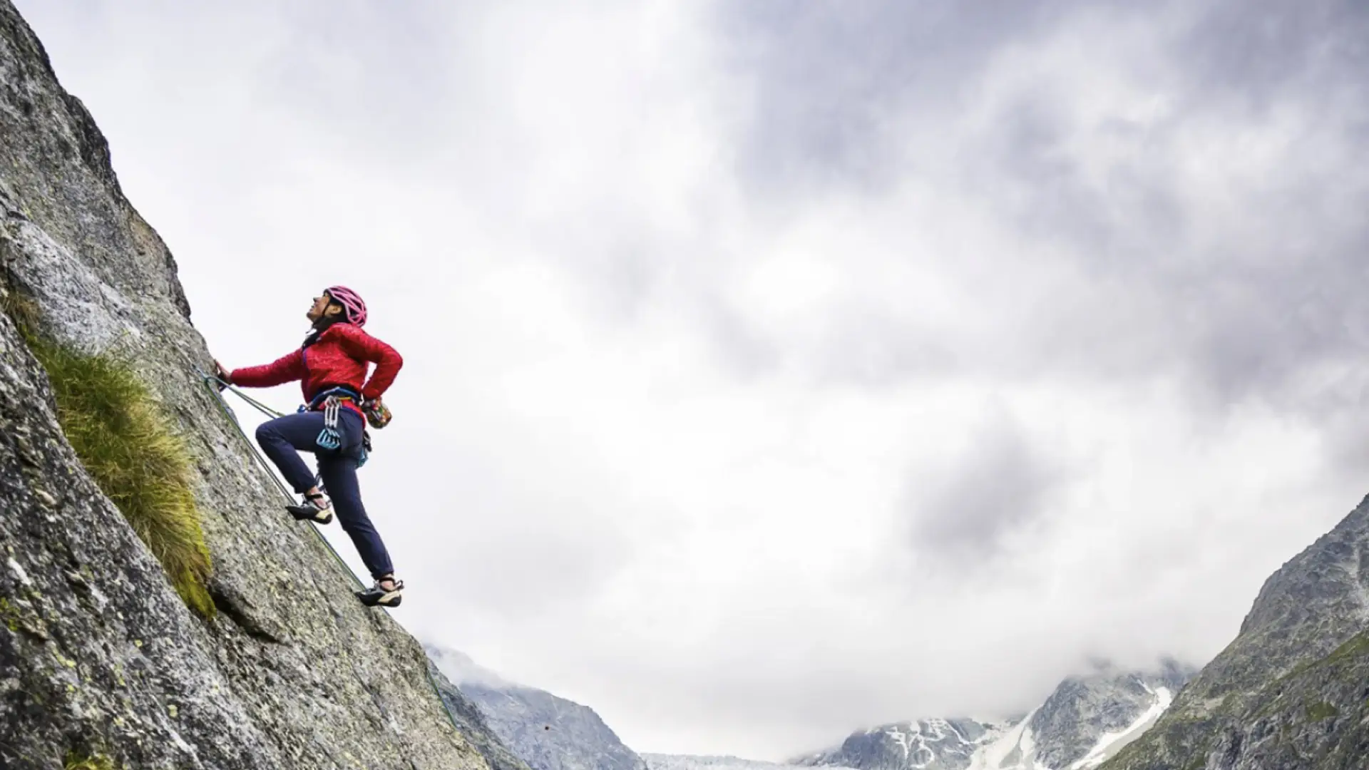 Woman in a red jacket and pink helmet rock climbing a granite slab, cloudy glaciated valley below