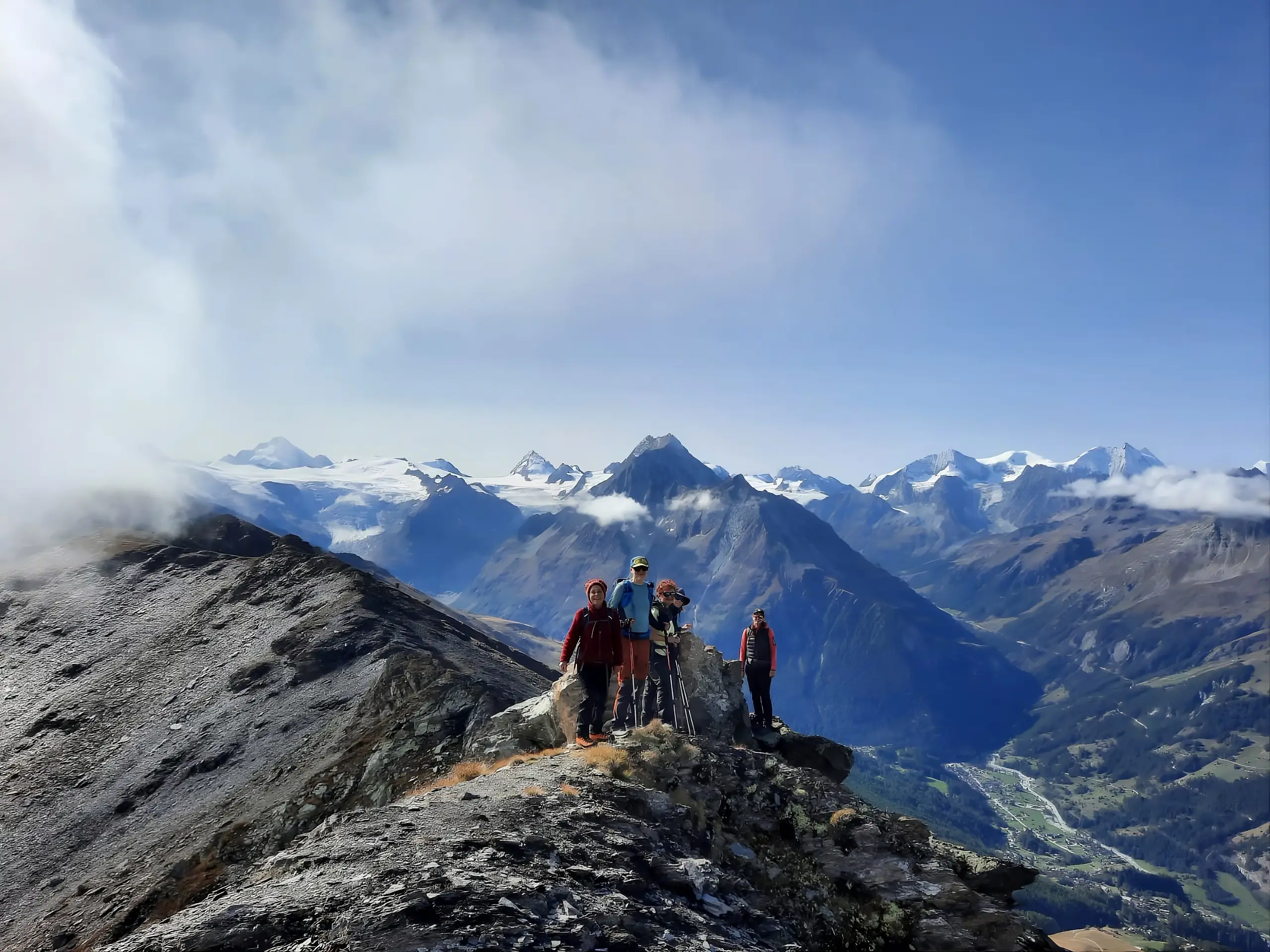 Fünf Wanderer mit Trekkingstöcken auf einem felsigen Grat, im Hintergrund erheben sich Gletscher und schneebedeckte Alpengipfel über einem tiefen Tal