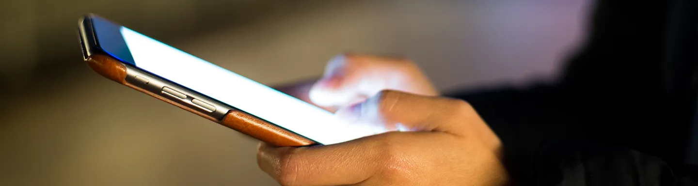 Close-up of a person’s hands holding a smartphone with a glowing screen in a dimly lit environment.