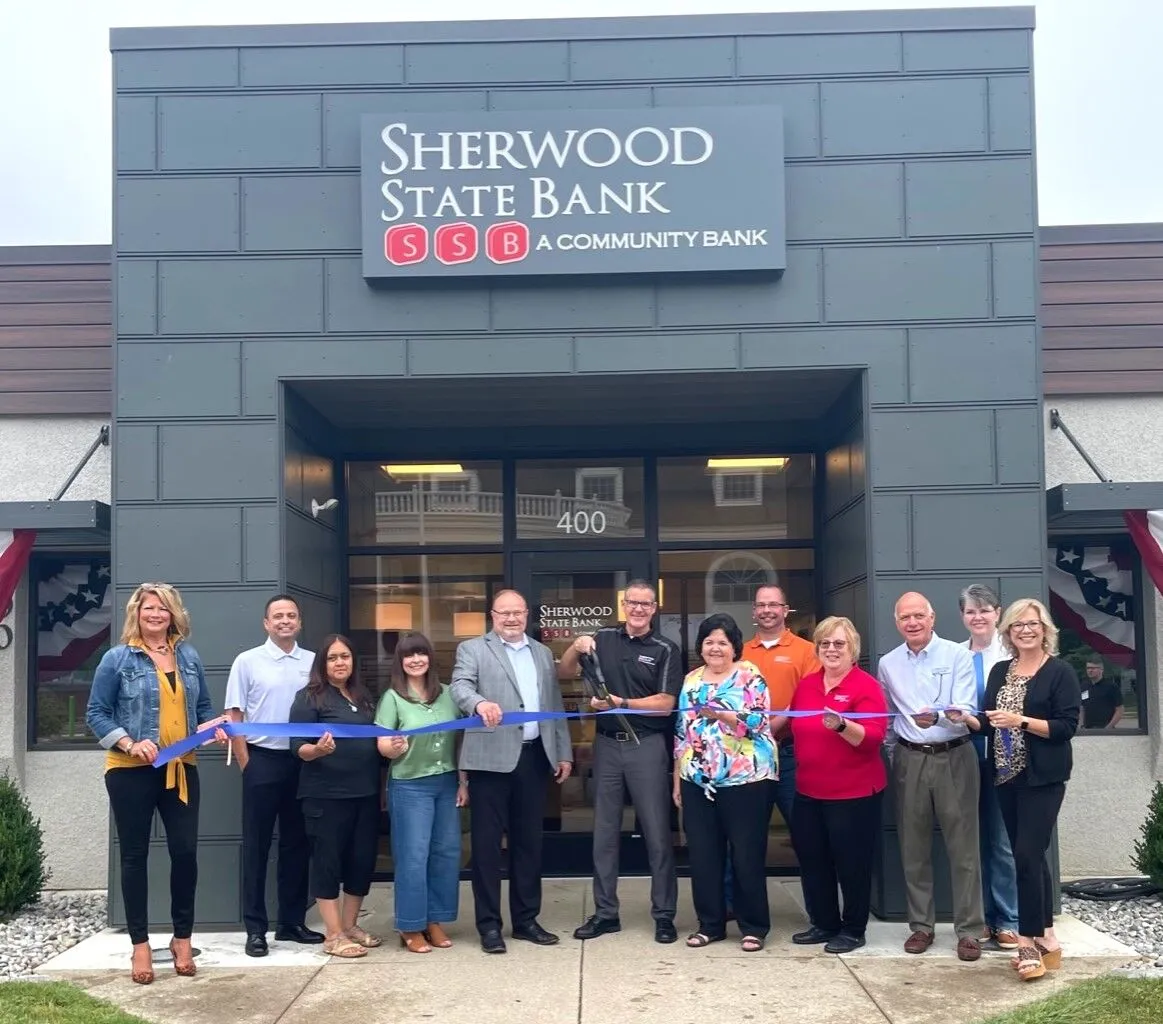 Group of twelve people smiling and holding a blue ribbon for a ribbon-cutting ceremony in front of Sherwood State Bank entrance.