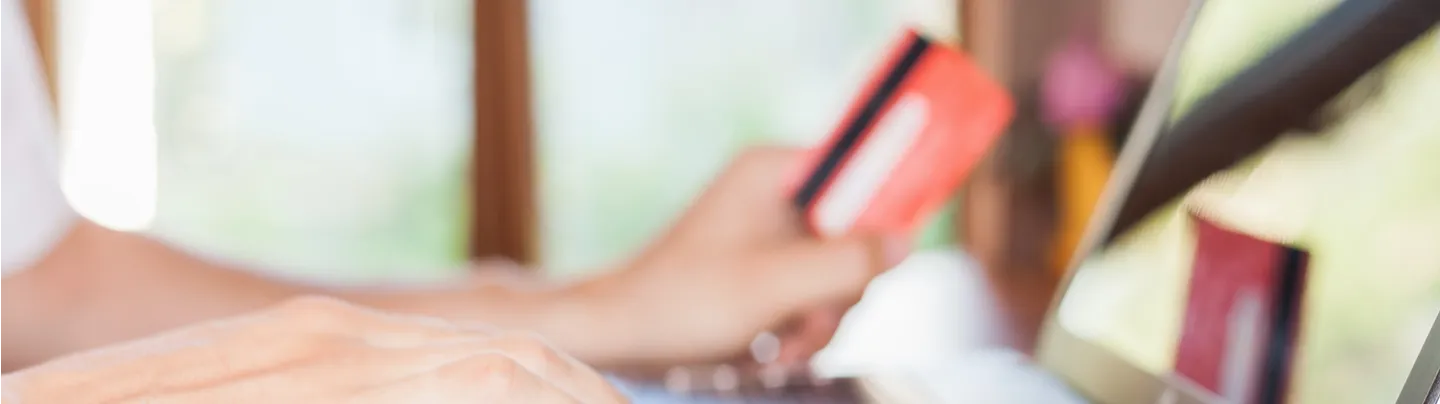 Person using a laptop with one hand typing on the keyboard and the other holding a red credit card.