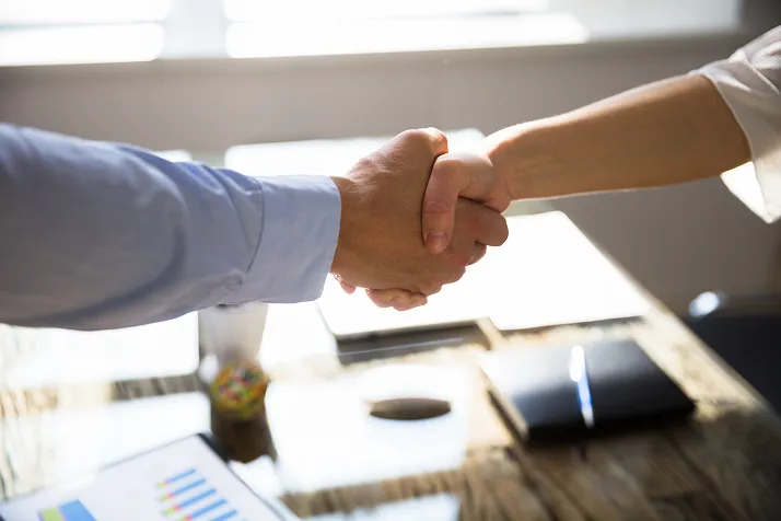 Two people shaking hands over a desk with documents and notebooks in a sunlit office.