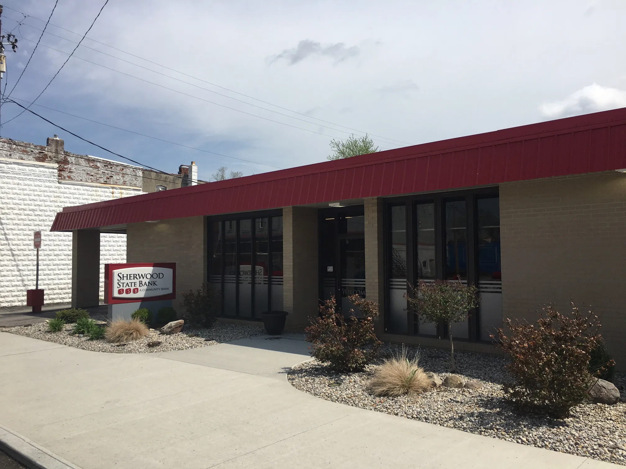Single-story Sherwood State Bank building with beige brick walls and a red roof canopy, surrounded by small bushes and gravel landscaping.
