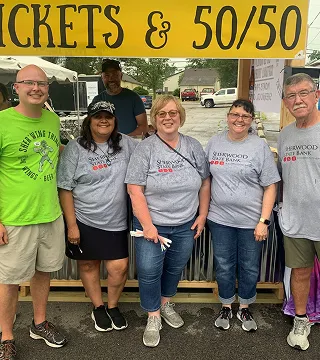 Five people standing smiling under a yellow tent banner that reads 'TICKETS & 50/50', wearing casual clothes and event T-shirts.