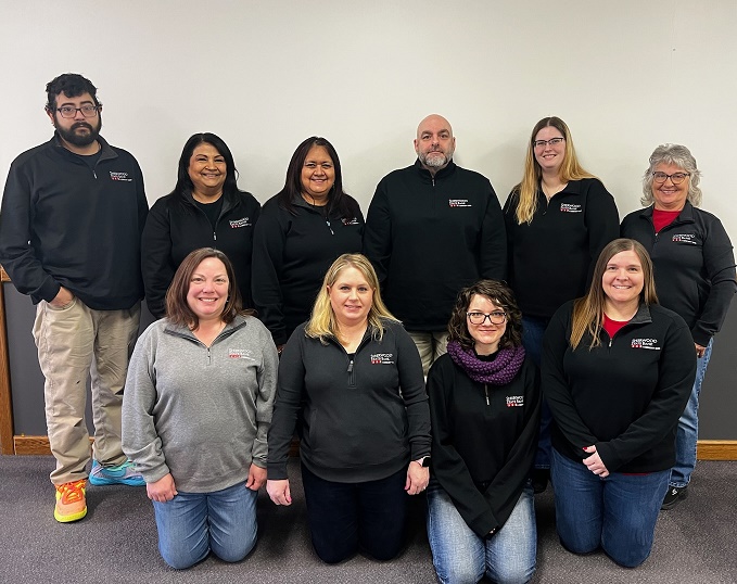 Four smiling people standing indoors in front of a beige wall, wearing Sherwood State Bank shirts in black, gray, and purple.