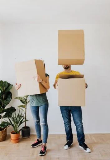Group of nine people including adults and children standing outdoors around a small white house structure with a gray roof in front of a large white building.
