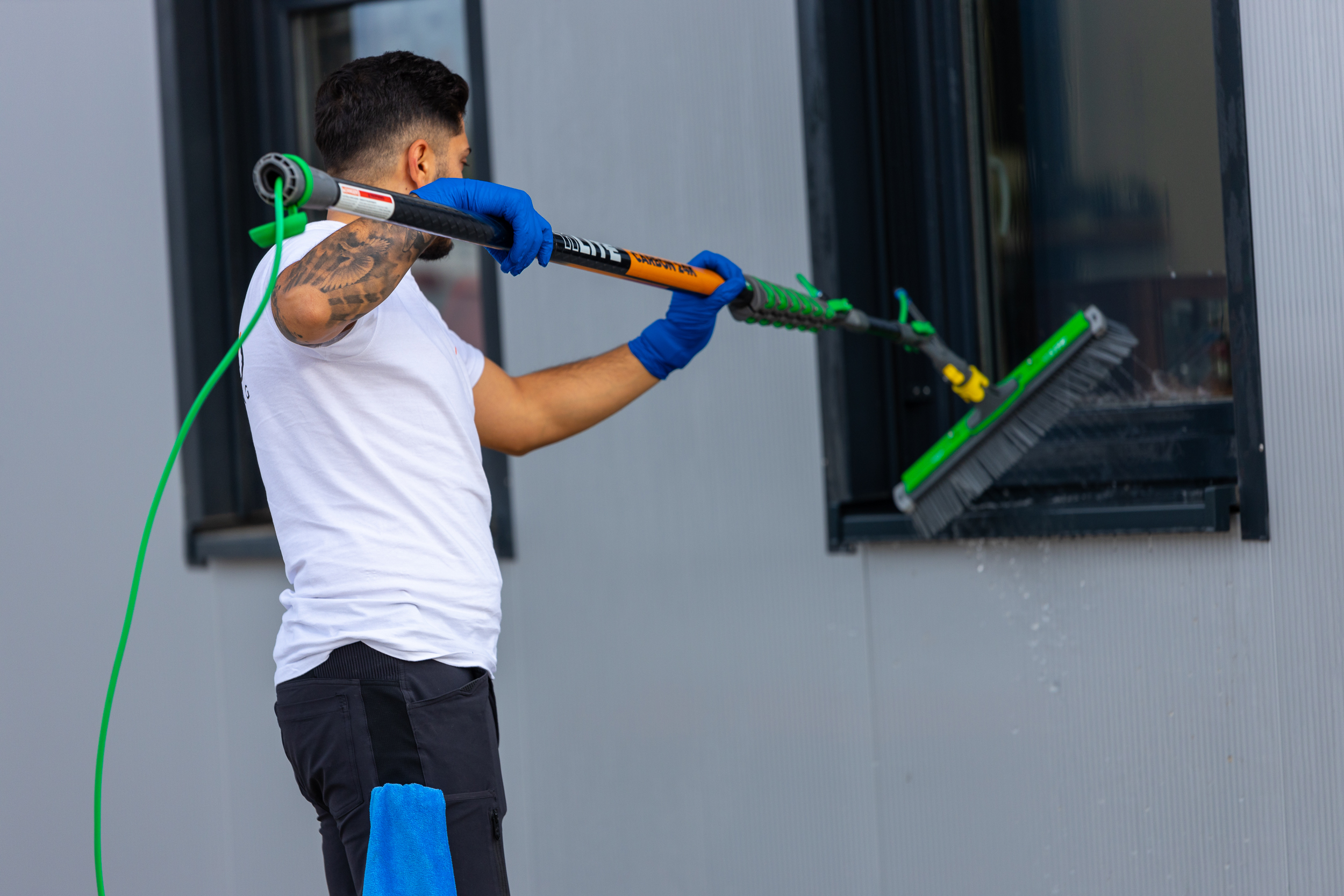 Man wearing blue gloves cleaning an outdoor window with a brush and hose.