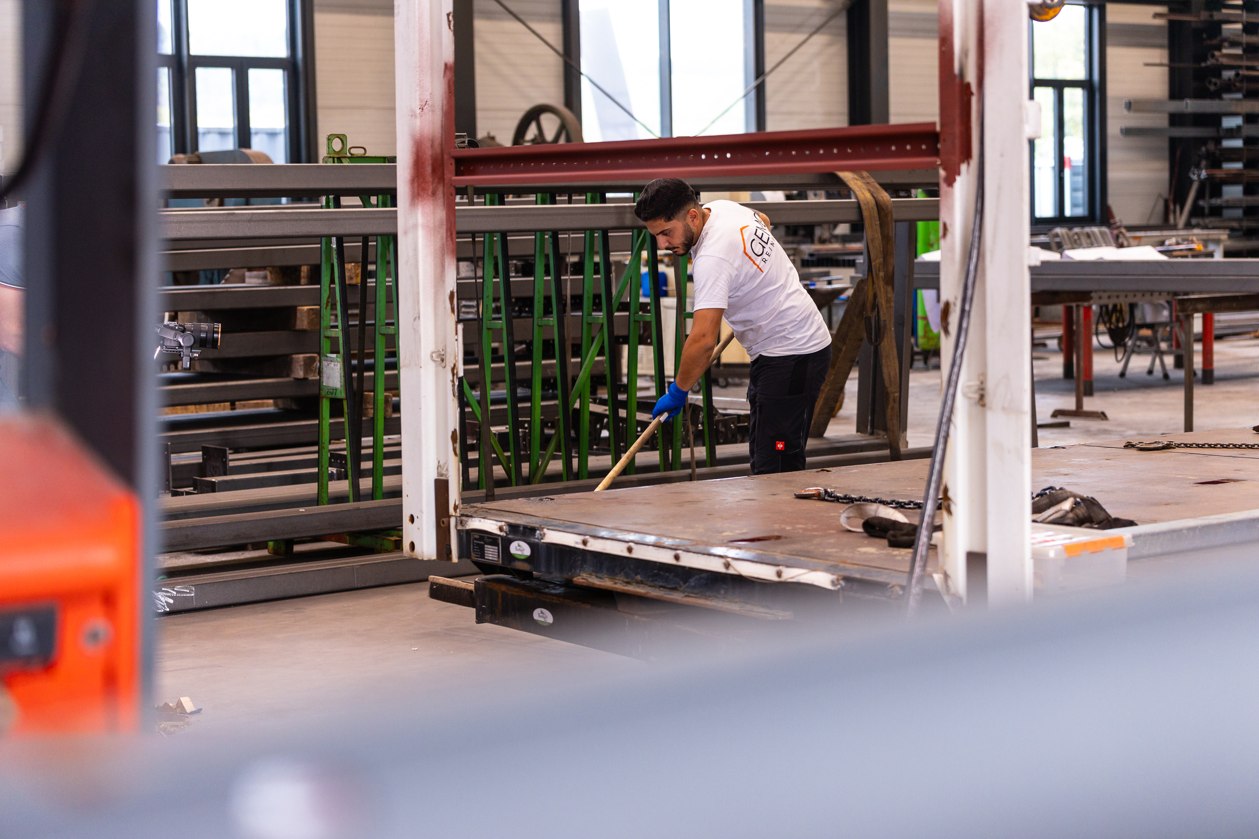 Worker in white shirt and blue gloves cleaning or sweeping a metal surface in an industrial workshop.