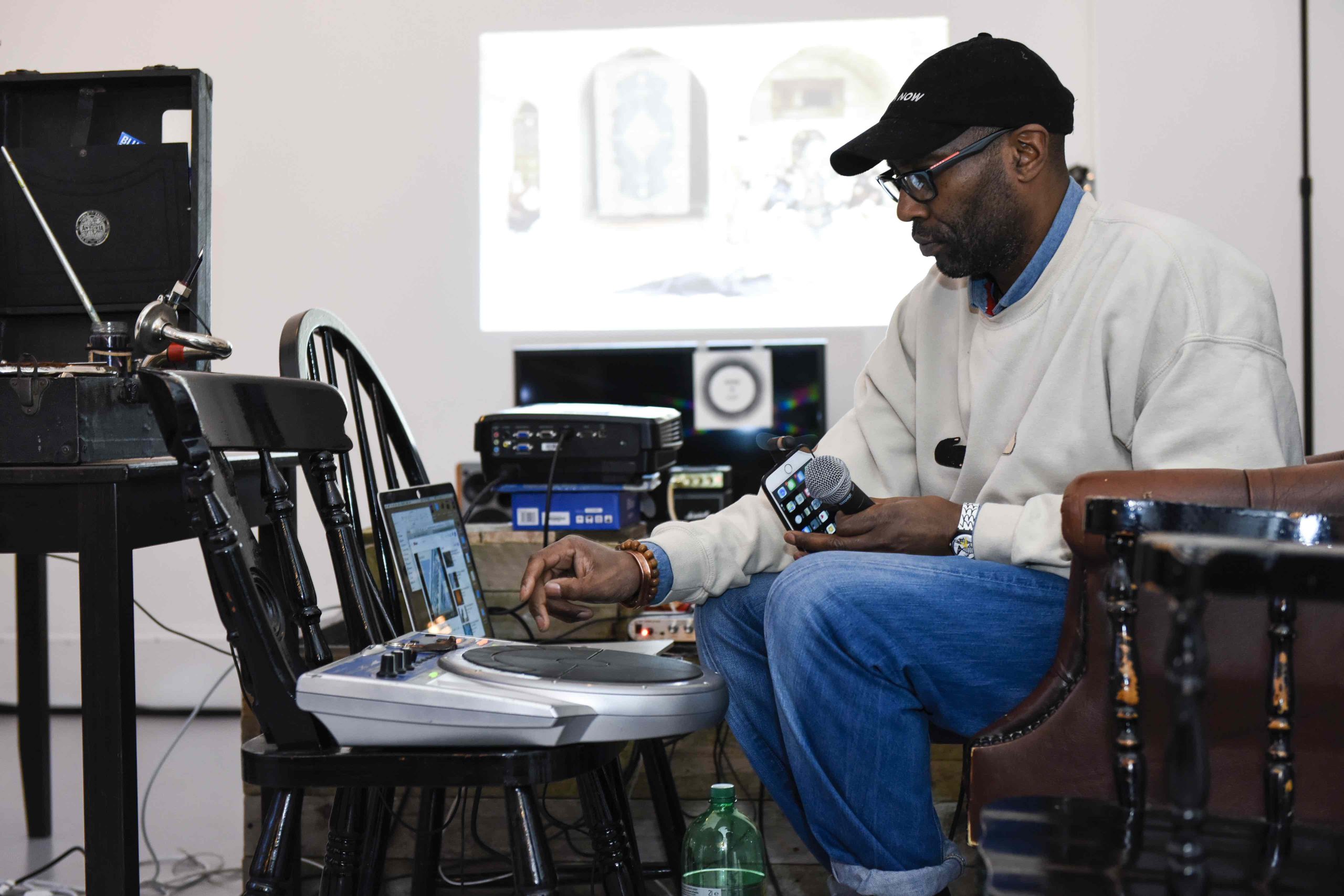 Man in black cap and glasses holding a microphone and phone, adjusting a digital drum pad on a chair with a laptop nearby.