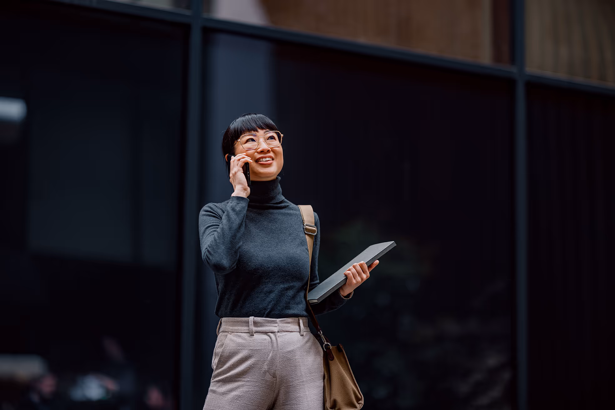 Smiling woman with glasses talking on a phone while holding a folder and a bag, standing outdoors.
