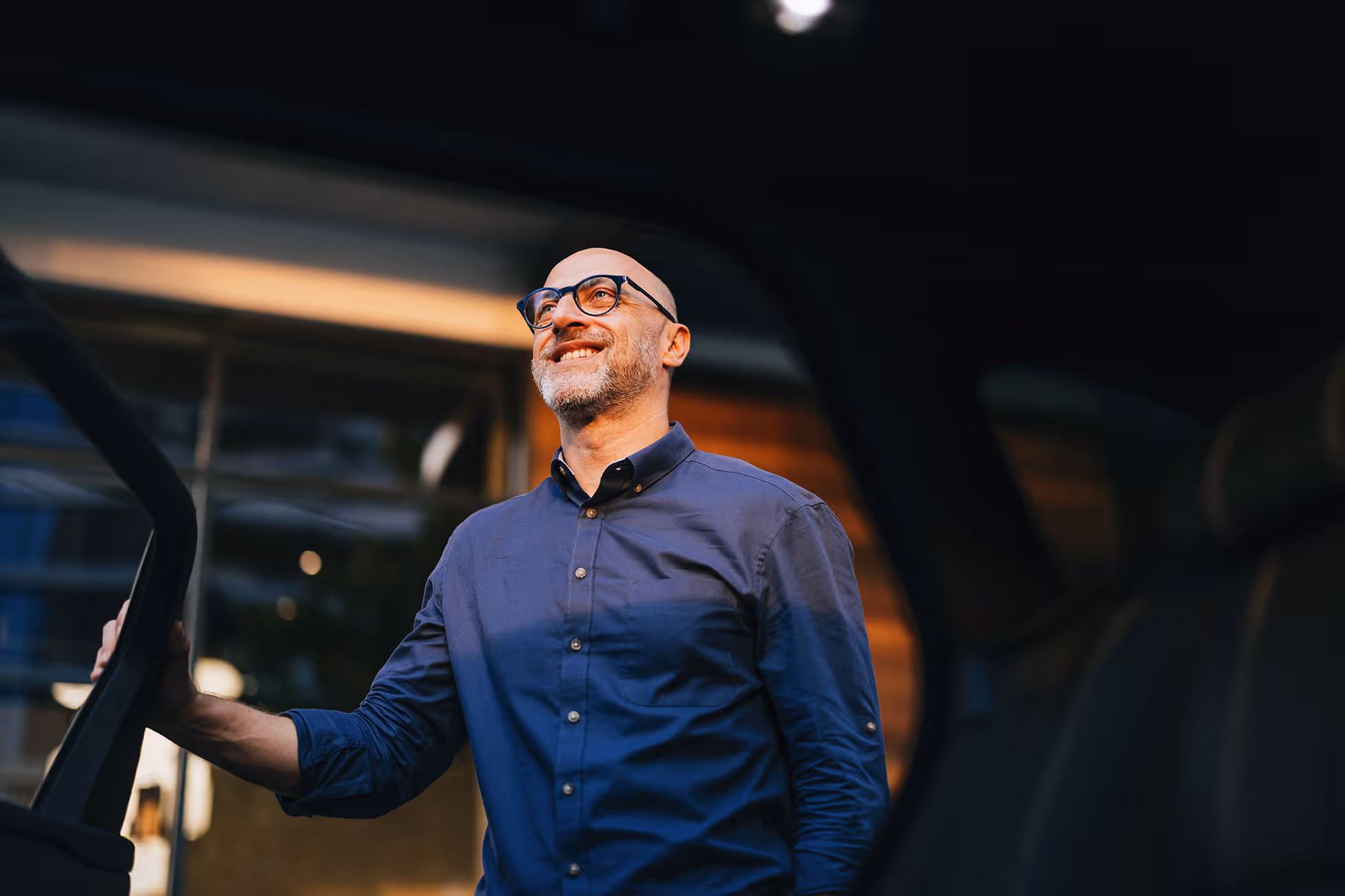 Smiling bald man with glasses wearing a blue button-up shirt standing outside a car holding the door open.