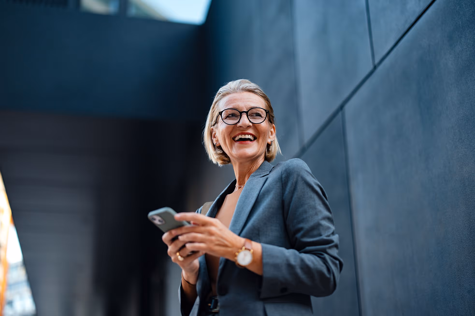 Smiling middle-aged woman in glasses and gray blazer holding a smartphone outdoors against a modern building.