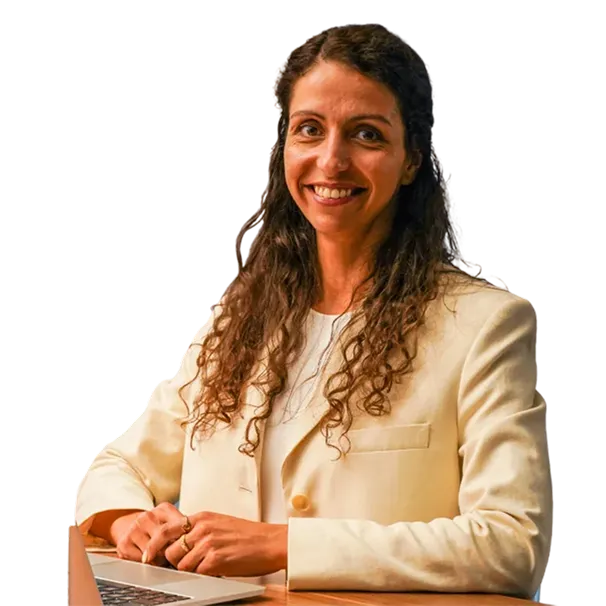 Smiling woman with long curly hair wearing a cream blazer, sitting at a desk with a laptop.