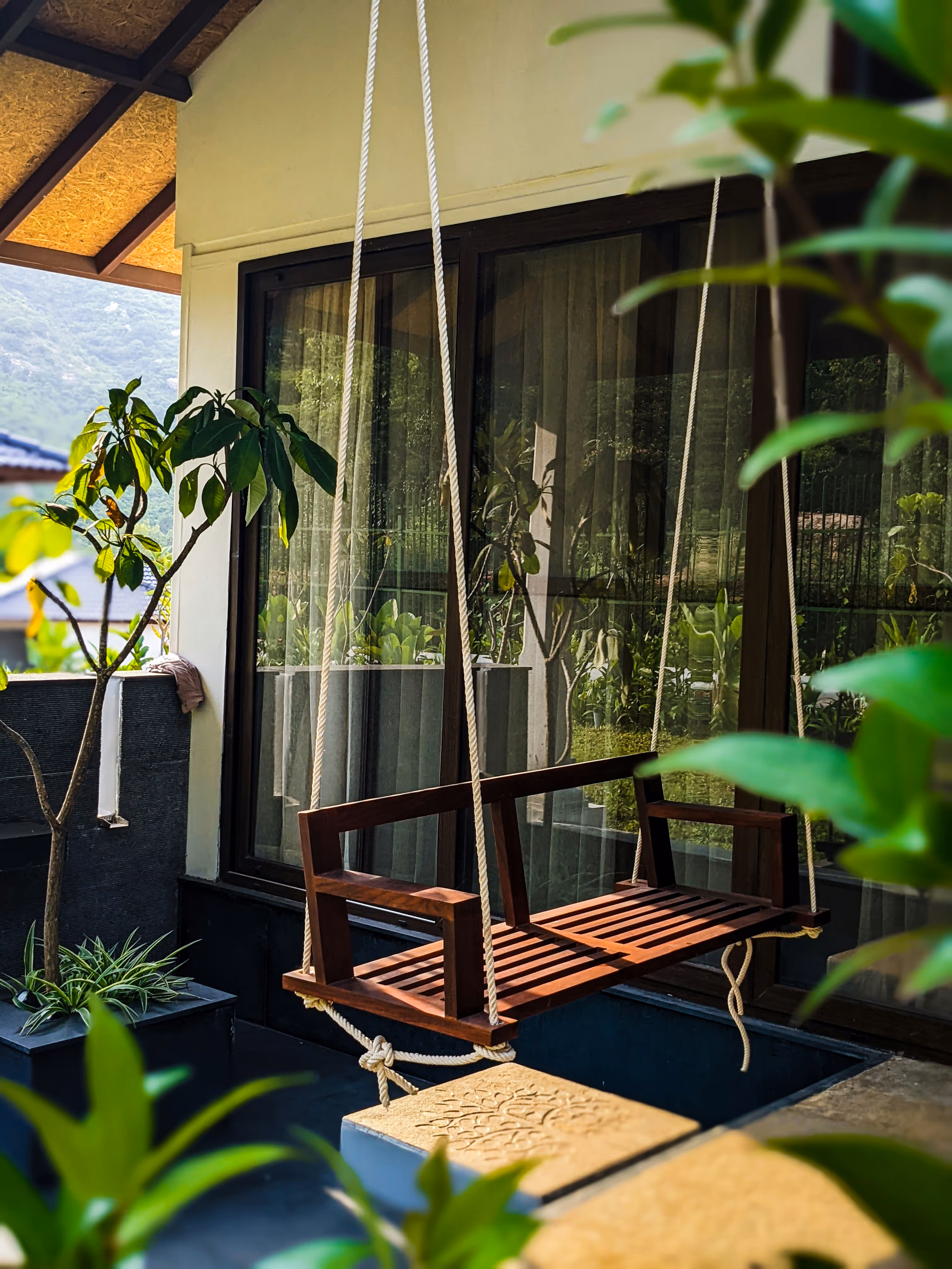 Wooden porch swing with rope support hanging in a garden patio area with plants and large glass windows reflecting greenery.