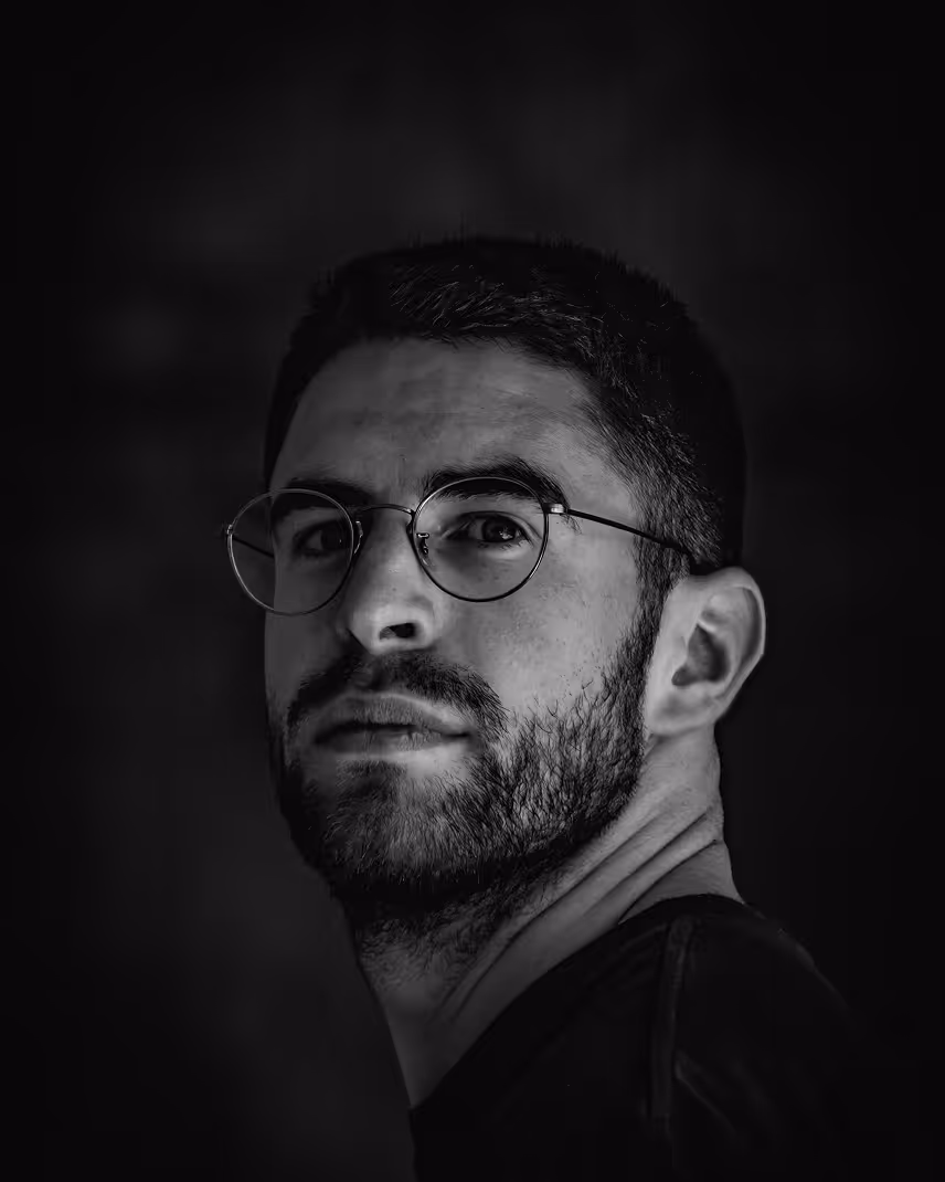 Black and white portrait of a man with glasses and a beard looking slightly to the side against a dark background.