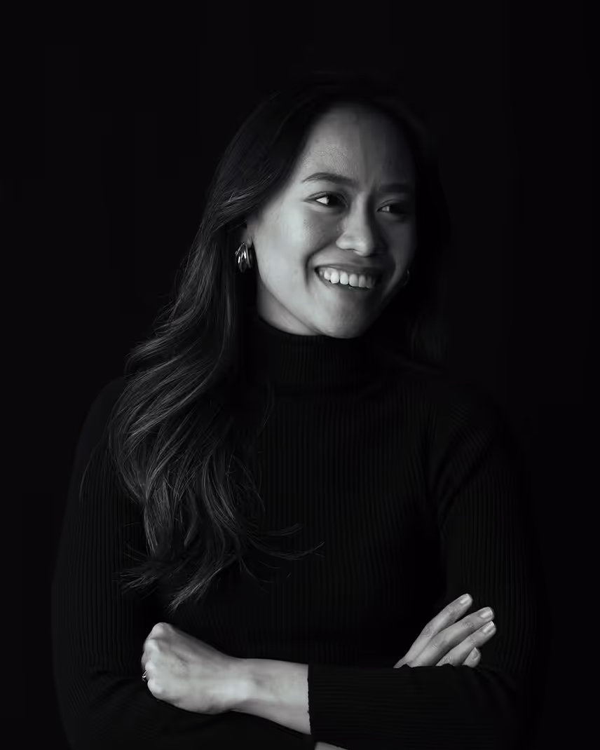 Smiling woman with long hair, wearing hoop earrings and a black turtleneck, arms crossed against a black background.