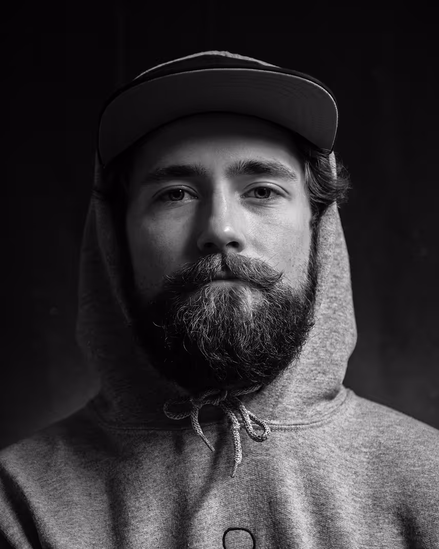 Black and white portrait of a man with a full beard and mustache wearing a hoodie and cap.