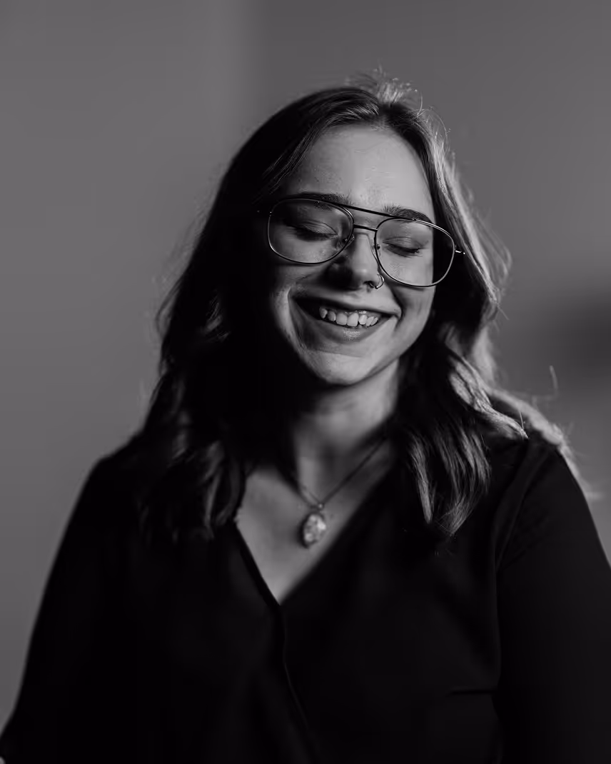 Smiling woman with glasses and a nose ring, wearing a necklace and a dark top, eyes closed in a black and white portrait.