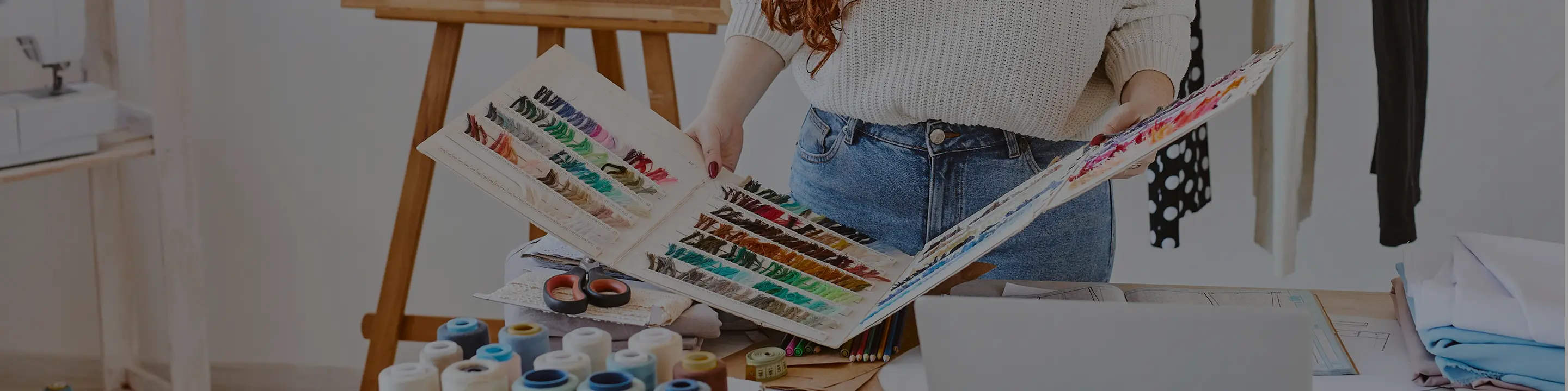 Person holding a large color swatch book with various thread samples over a cluttered craft table with scissors, spools of thread, and fabric.