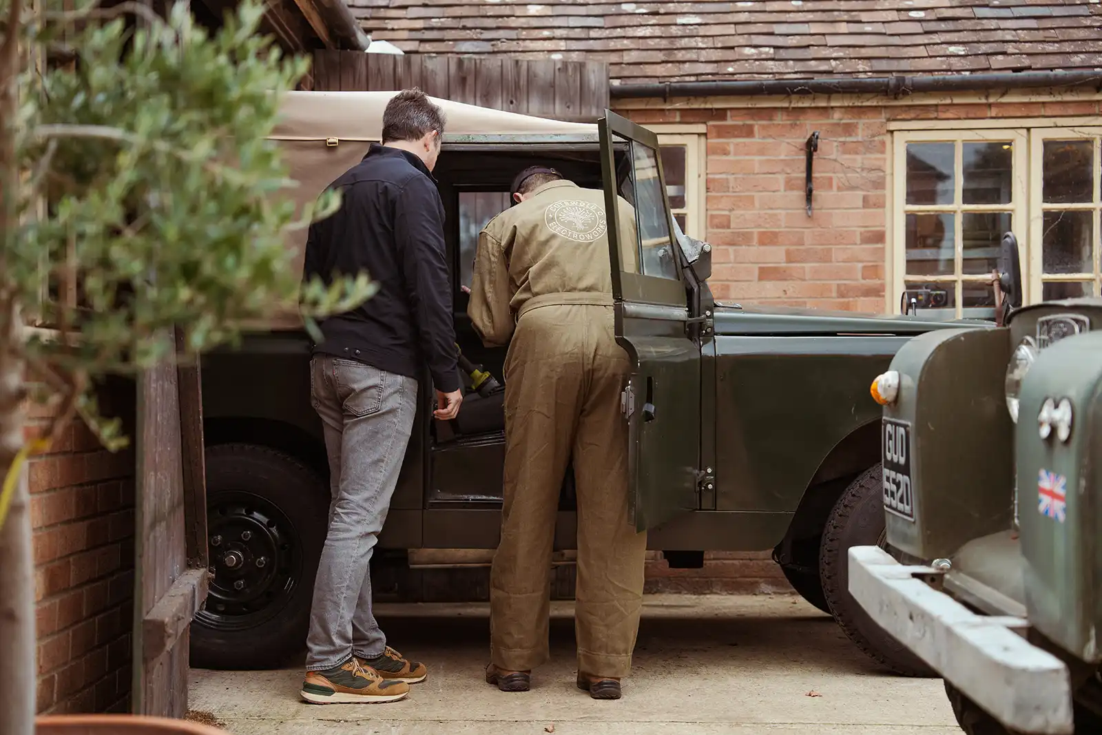 Two people standing near vintage Land Rover in brick building courtyard