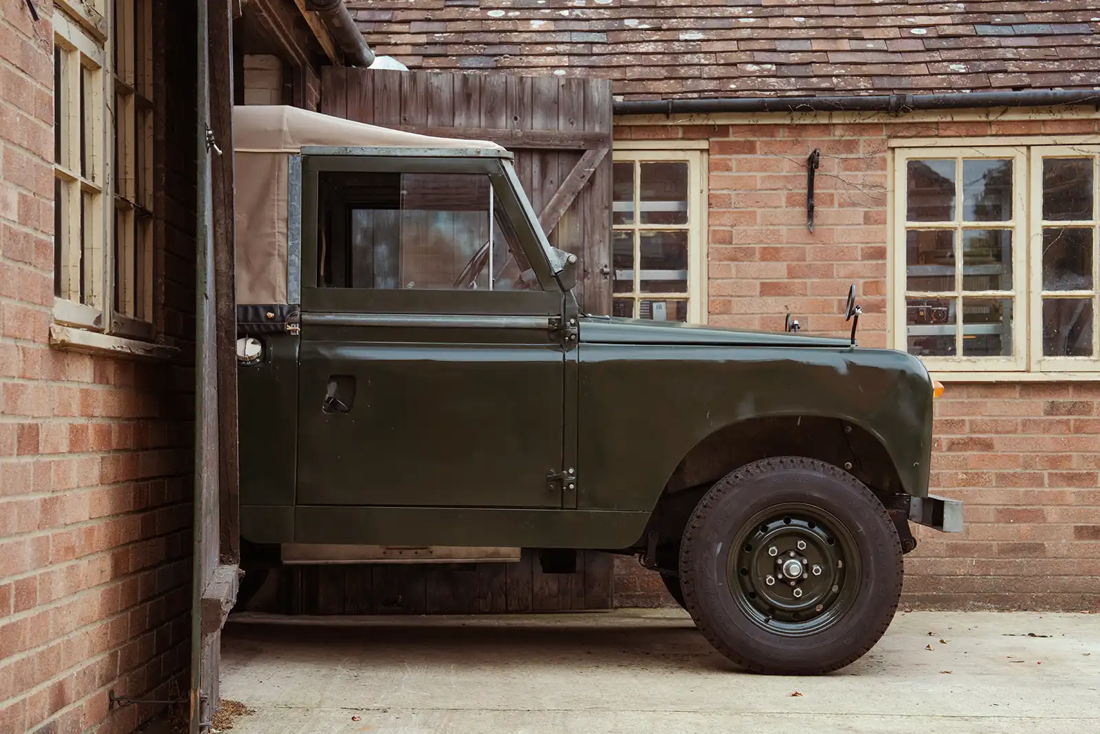 Vintage dark green Land Rover parked beside brick building with wooden barn