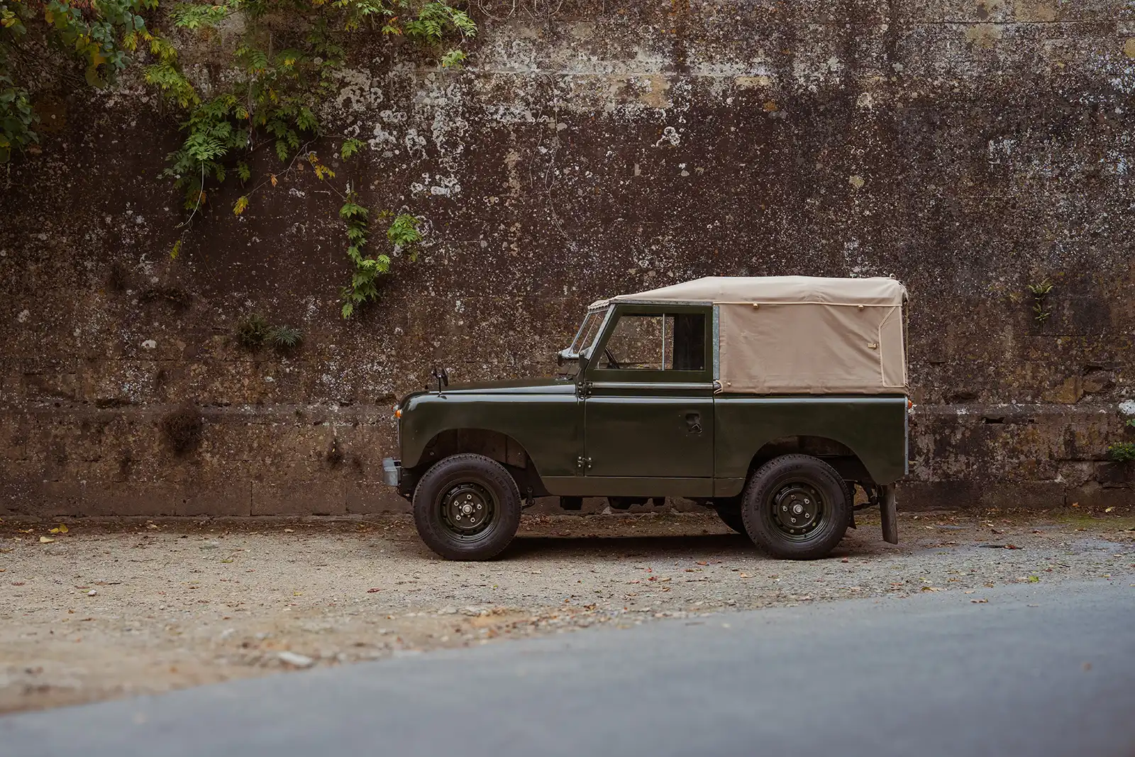 Classic green Land Rover with white soft top parked against weathered stone wall