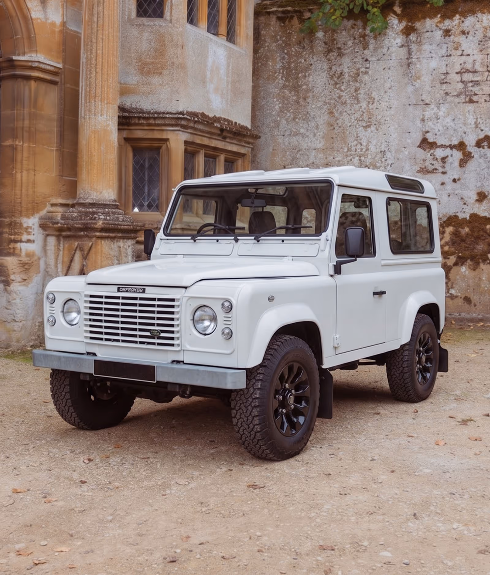 White Land Rover Defender parked on gravel in front of an old stone building.