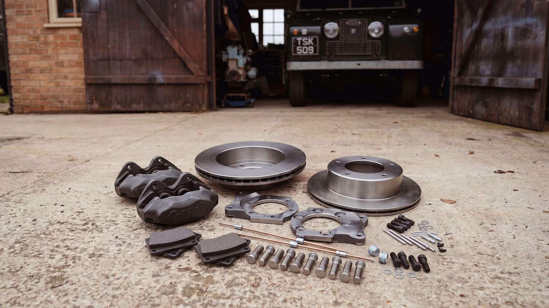 Car brake kit with rotors, calipers, pads, and bolts arranged on a concrete floor in front of a garage.