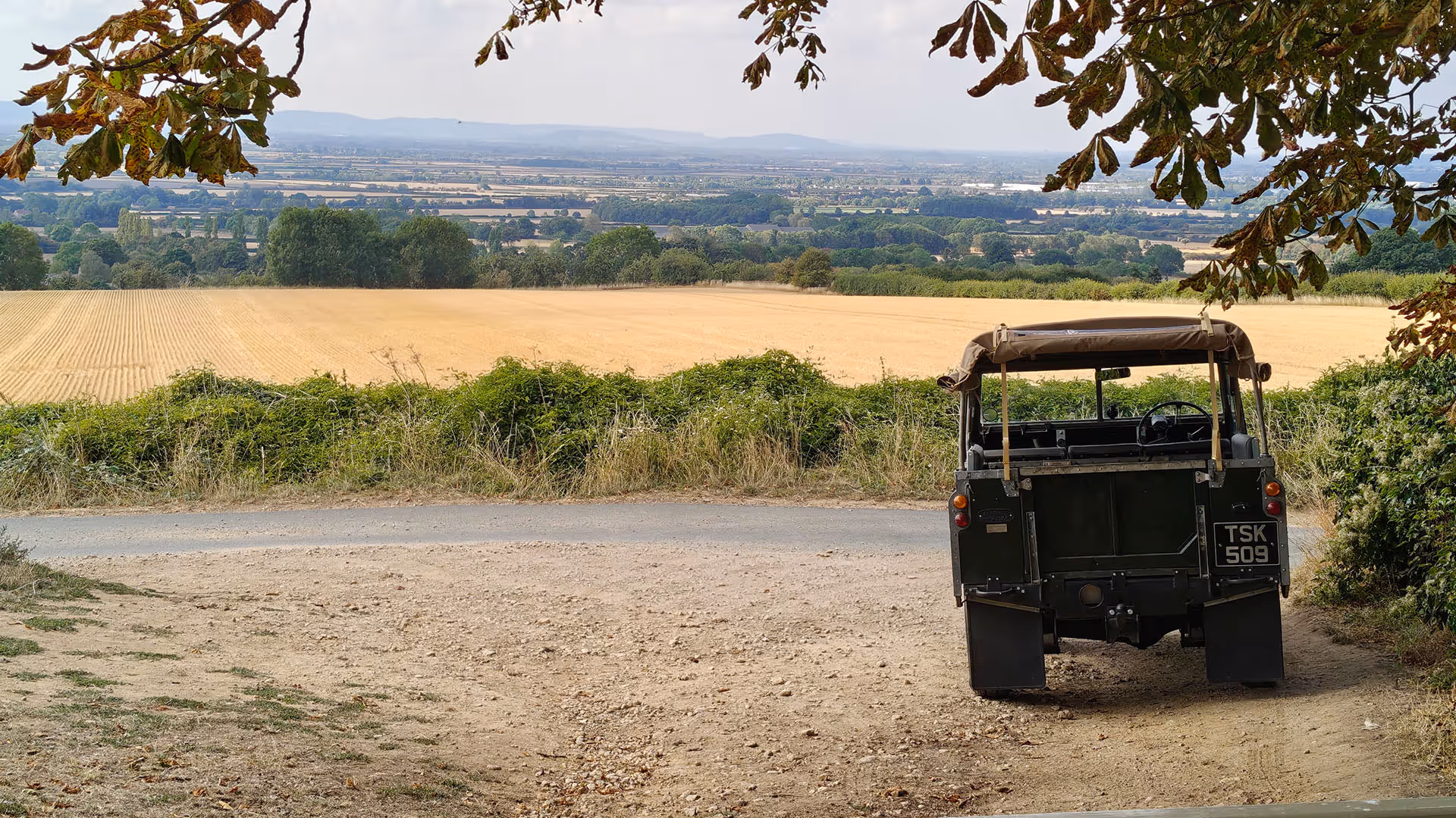 Rear view of a vintage green off-road vehicle parked on a dirt path overlooking golden fields and distant hills.