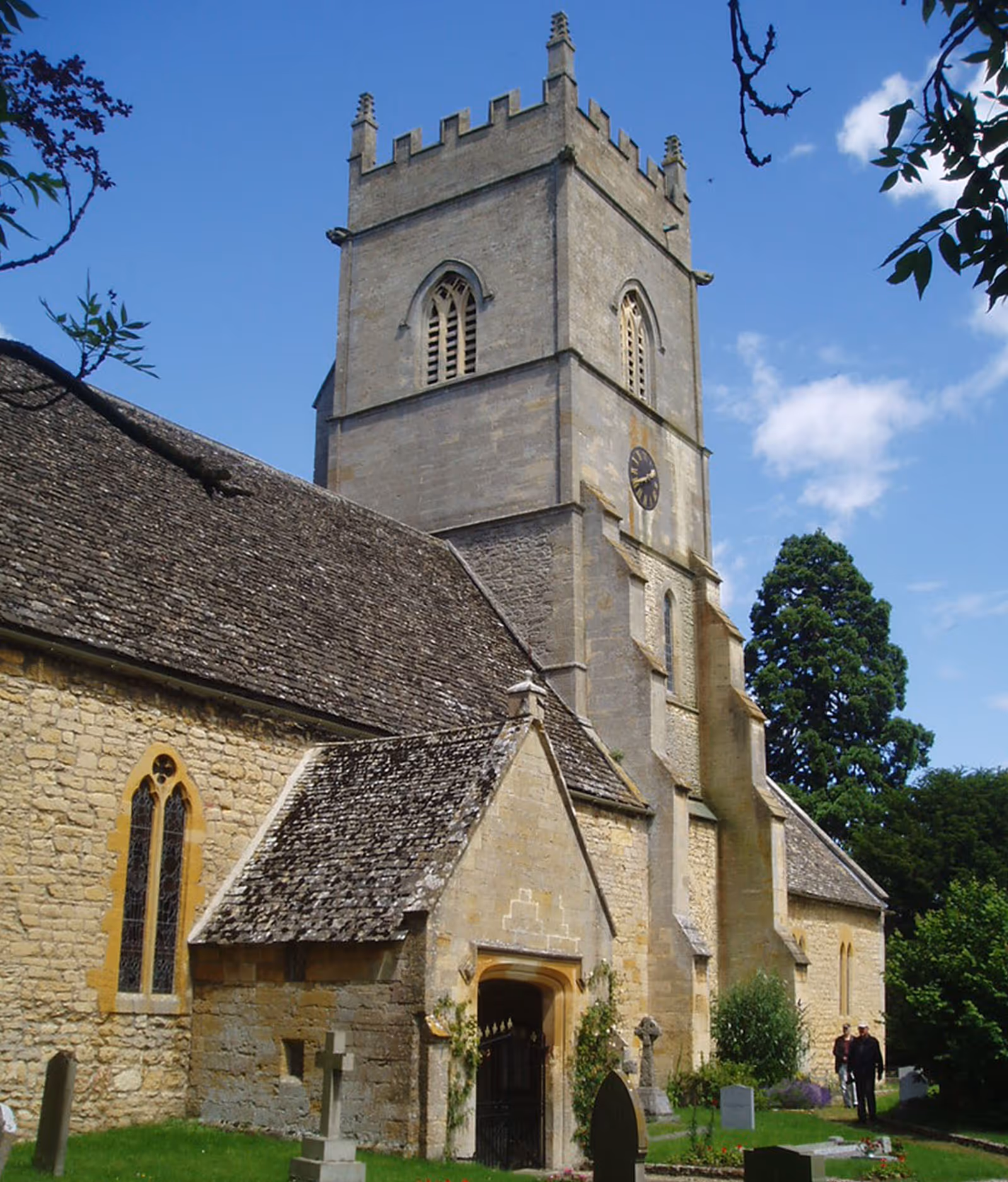 Stone church with a square clock tower, arched windows, and surrounding graveyard under a blue sky.