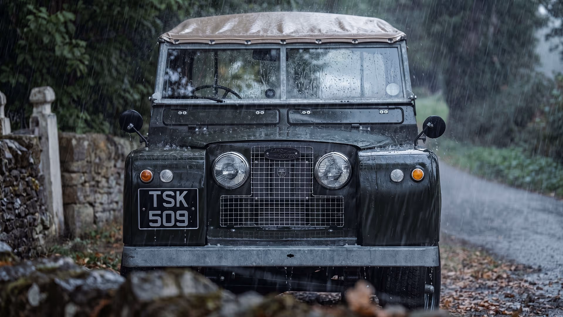Front view of a vintage Land Rover with a soft top driving on a rainy country road.