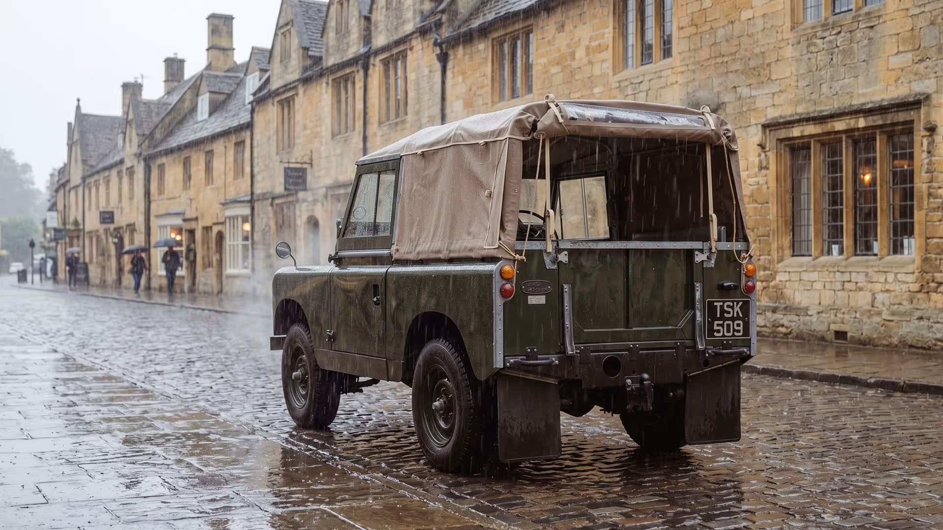Vintage green Land Rover with beige canvas top parked on a wet cobblestone street in rainy weather.