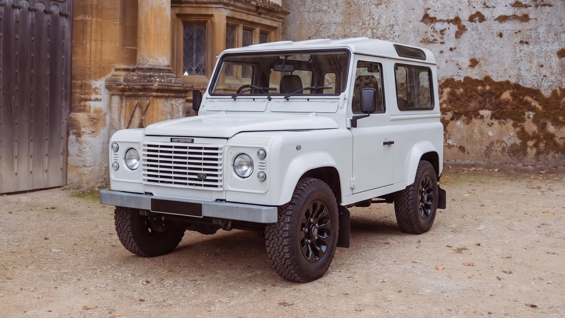 White Land Rover Defender parked on gravel in front of an old stone building.