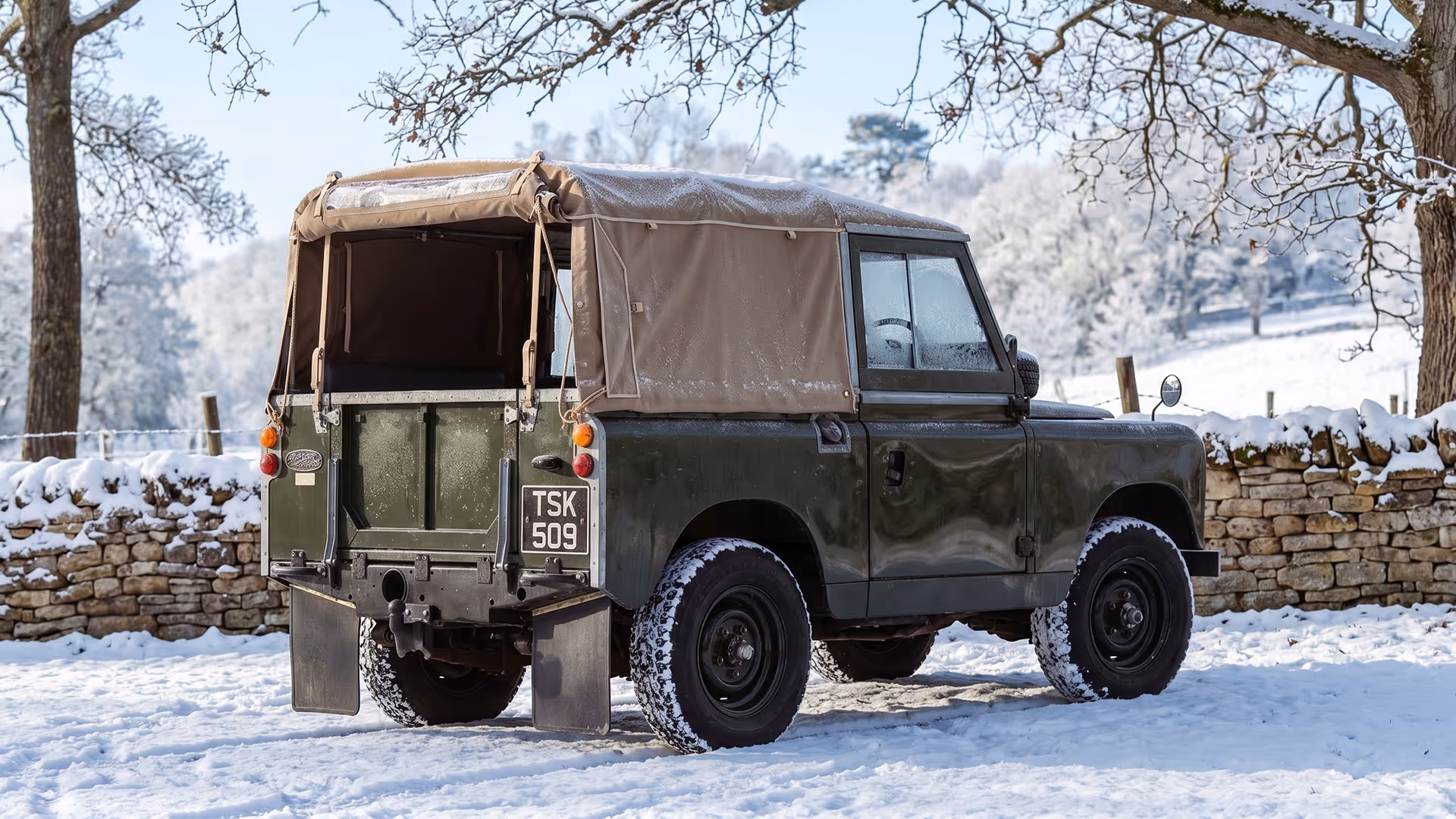 Vintage green Land Rover with beige soft top parked on snow-covered ground near stone wall and leafless trees.