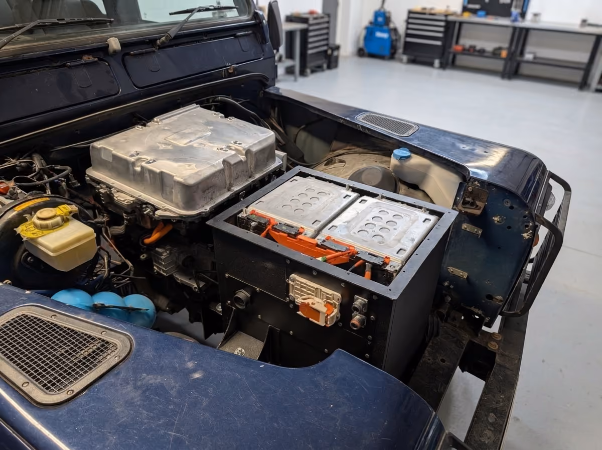 Close-up of an electric vehicle's engine compartment showing battery packs and power electronics inside a blue vehicle in a workshop setting.