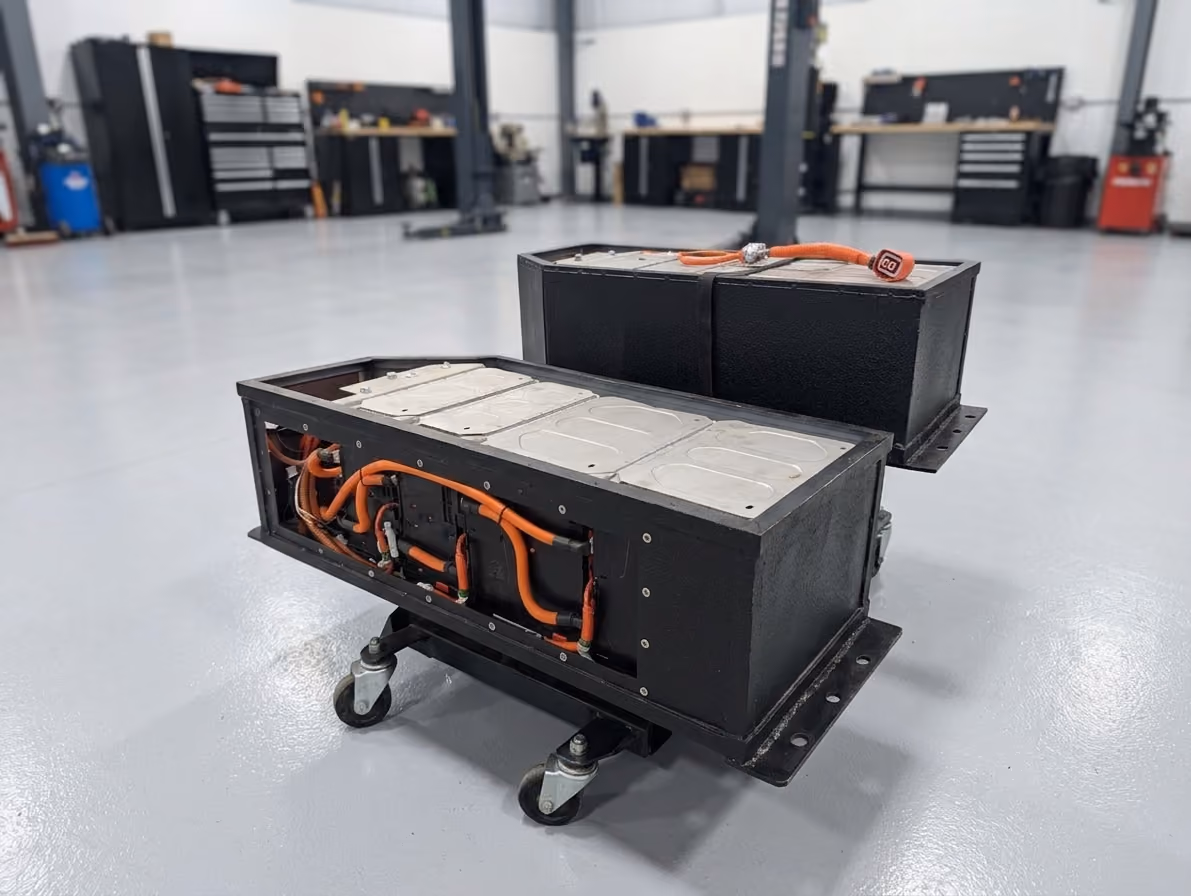 Large electric vehicle battery pack with multiple rectangular cells connected by orange cables.