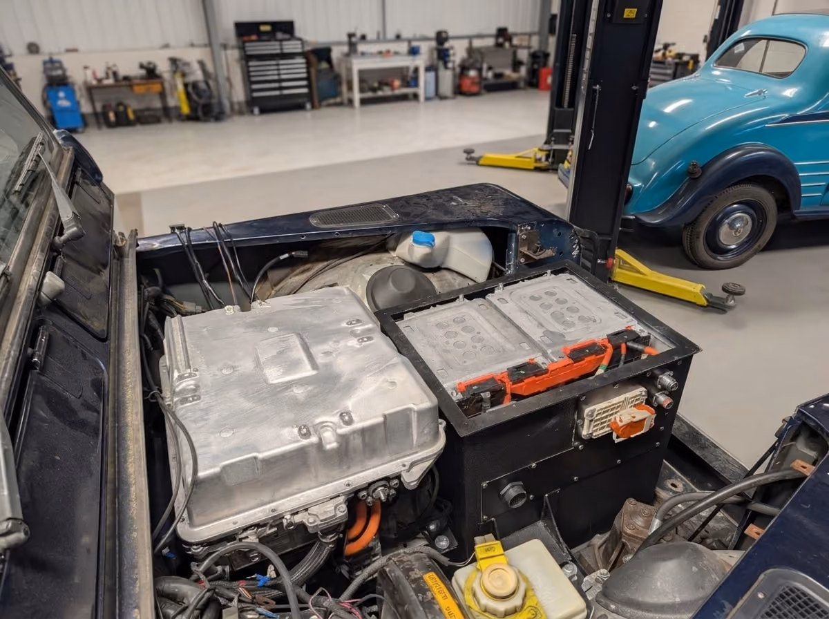 Close-up of an electric vehicle's engine compartment showing a large battery pack, a silver metal component, various cables, and fluid reservoirs, with a blue vintage car and workshop tools visible in the background.