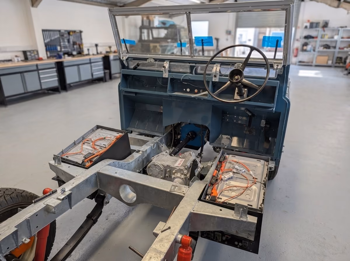 Partially assembled vehicle chassis with exposed electric batteries, wiring, and drivetrain components inside a spacious workshop.