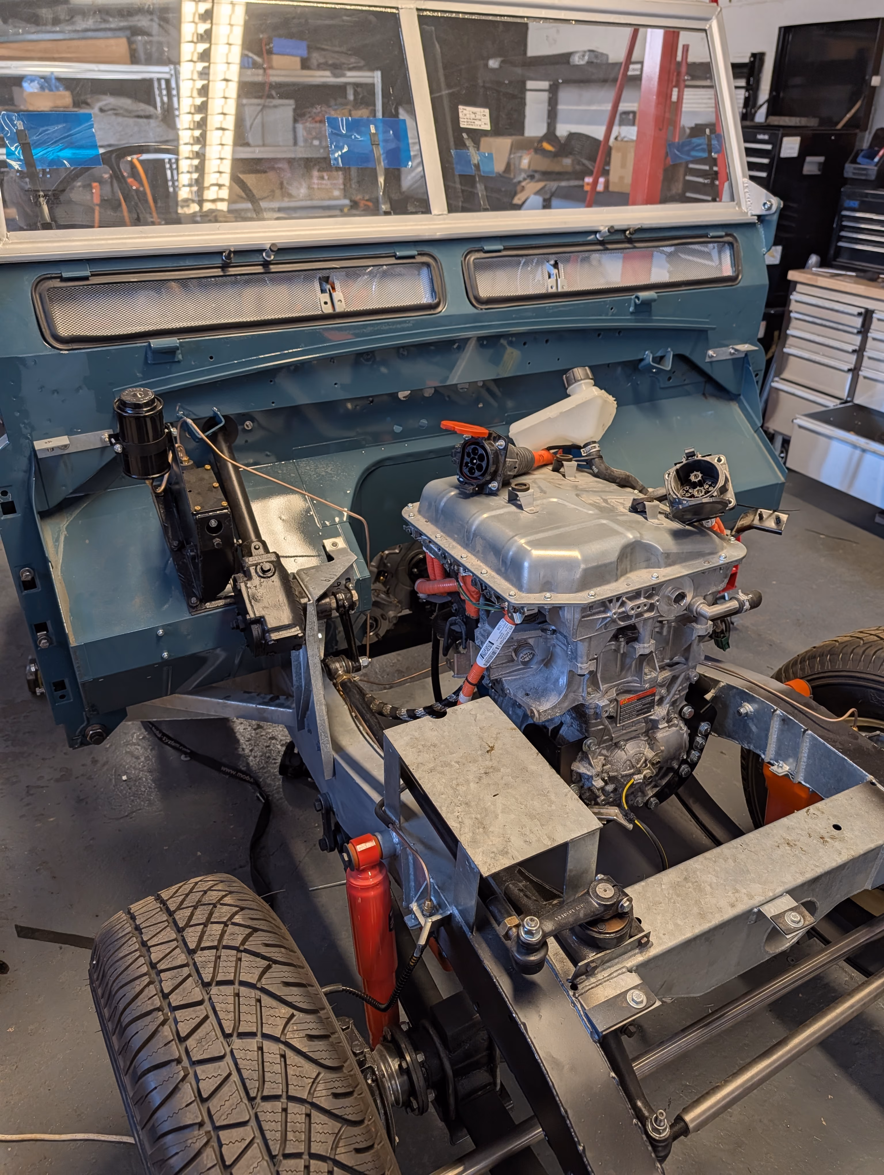 Partially assembled vehicle chassis with an engine mounted, front suspension components, and a windshield frame in a workshop with tools and shelves in the background.