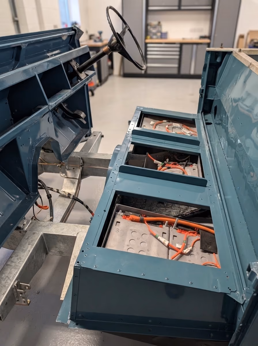 Interior view of a partially assembled electric vehicle chassis with visible battery compartments containing orange and black wiring, a steering wheel, and a workshop background.